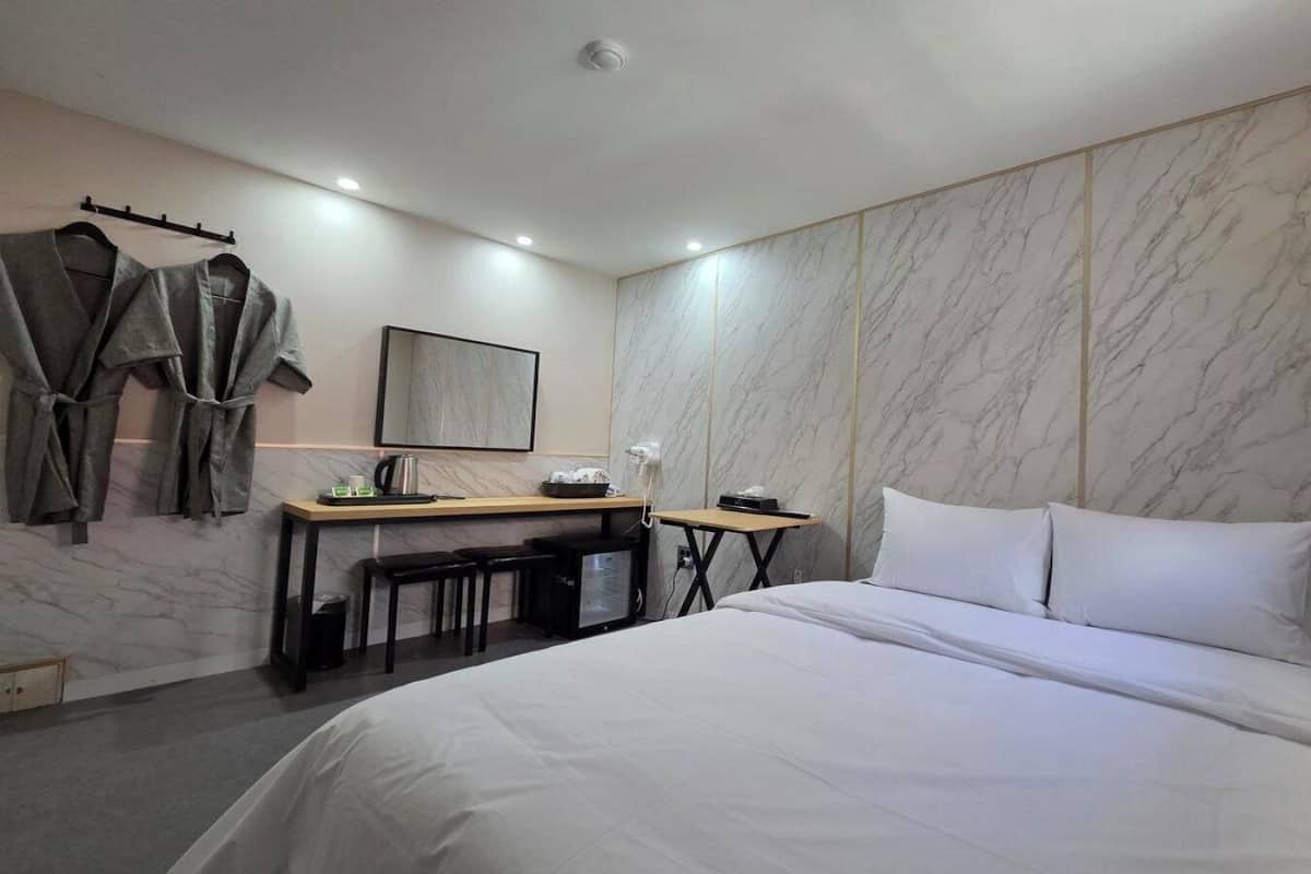 Une chambre d’hôtel comprenant un lit, un bureau avec une chaise, une petite table, un miroir et des peignoirs suspendus à un porte-manteau.