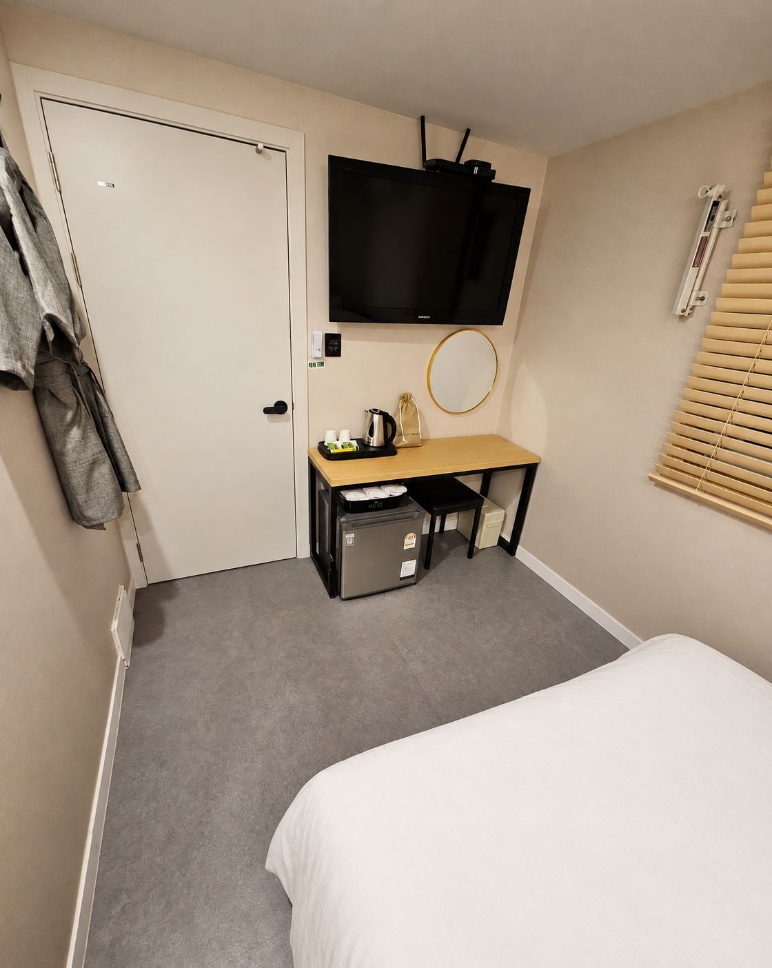 Quarto de hotel com cama, escrivaninha com televisão, mesinha com chaleira e janela com persianas.