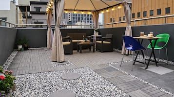 Ein Außenbereich mit einer überdachten Sitzgruppe, einem Tisch mit Stühlen und Blick auf die Stadtlandschaft im Hintergrund.