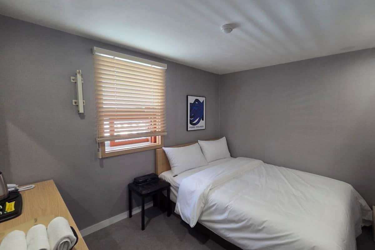 Une chambre à coucher comprenant un lit, un bureau avec un téléphone, une chaise, une fenêtre avec des stores et un téléviseur fixé au mur.