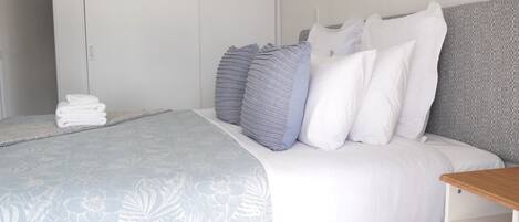 A neatly made bed with a floral-patterned bedspread, a stack of white towels, and a grey headboard.