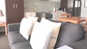A modern living room with a grey sofa, a wooden dining table, and a kitchen in the background. - 2 Bedroom Beach Frontage Apartment (Paihia)
