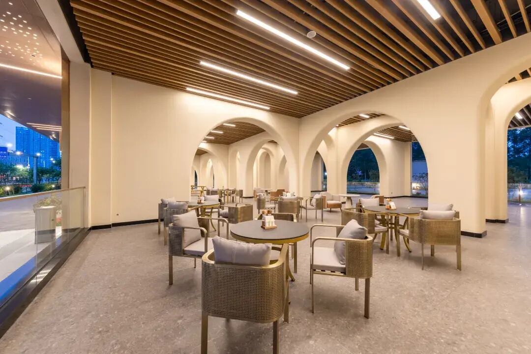 A spacious indoor area with multiple tables and chairs, a wooden ceiling with linear lights, and a view of the cityscape through large windows.