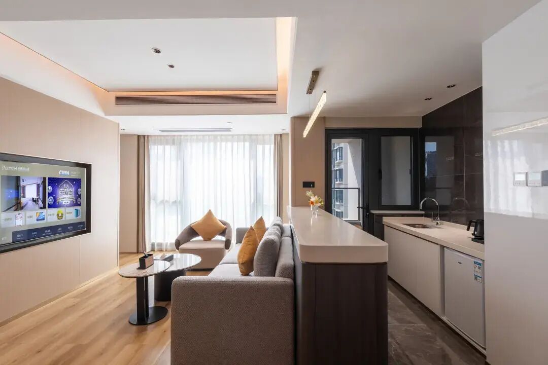 A modern hotel room with a sofa, a small table, a TV, a kitchen area, and a large window.