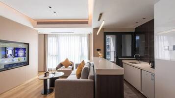 A modern hotel room with a sofa, a small table, a TV, a kitchen area, and a large window.