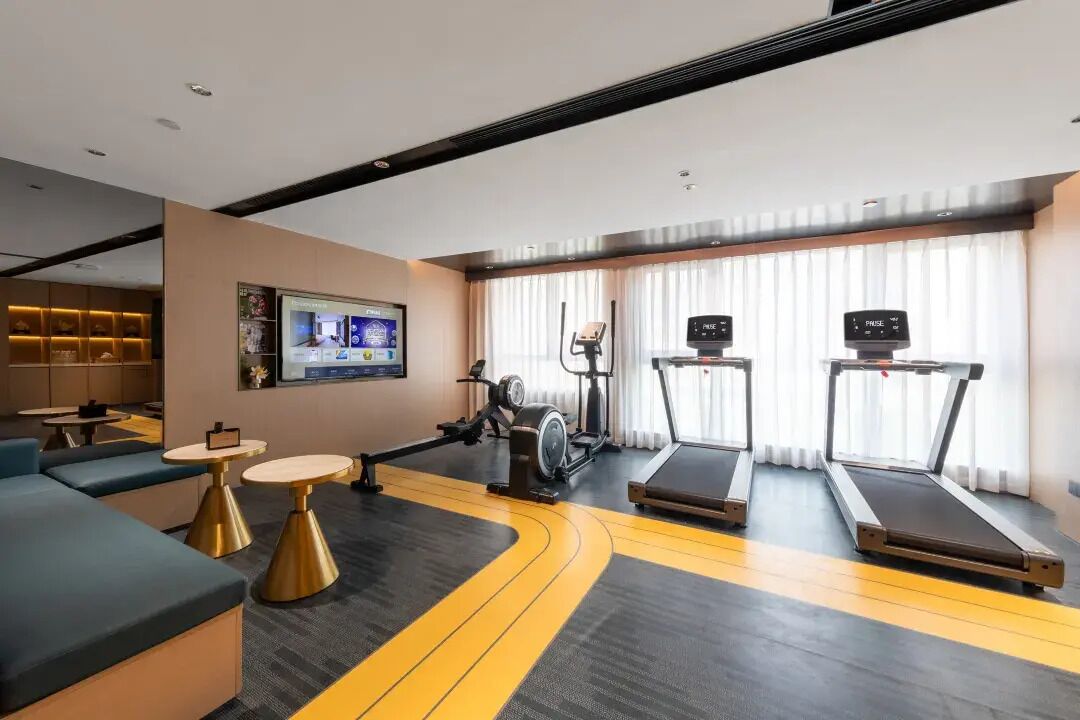 A modern hotel room with a gym area, including treadmills and exercise equipment, a TV, and seating with tables.