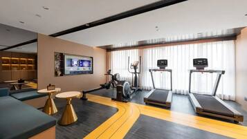 A modern hotel room with a gym area, including treadmills and exercise equipment, a TV, and seating with tables.