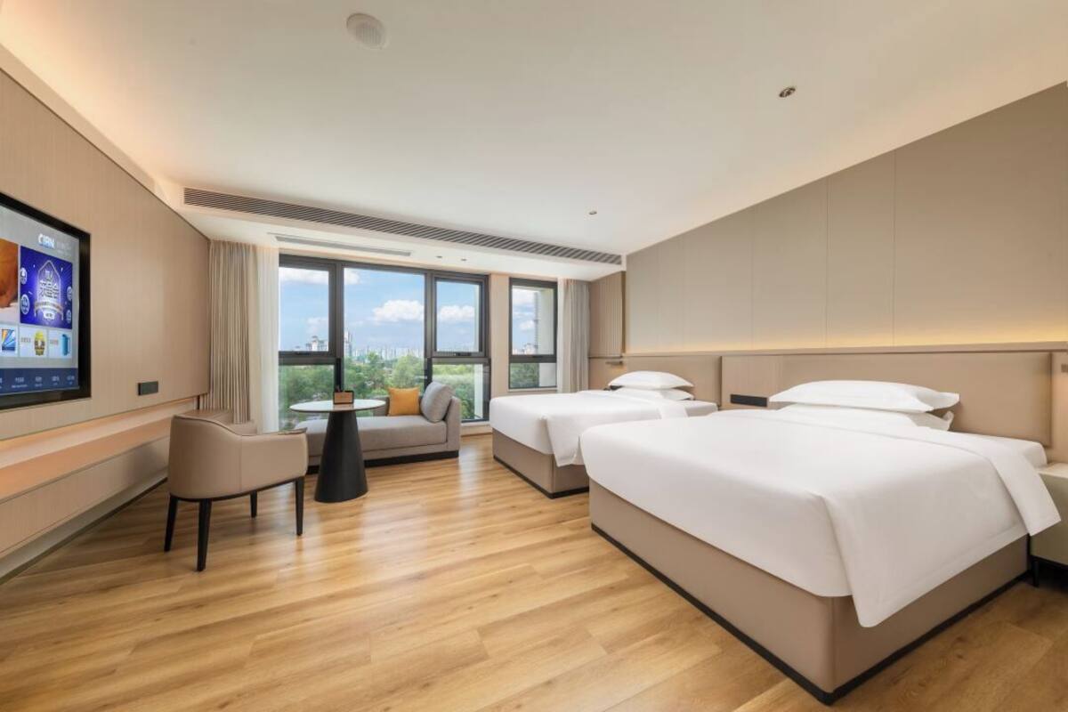 Habitación de hotel con televisor grande, sofá, una mesita, dos camas y vista a la ciudad.