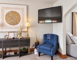 A living room with a blue armchair, a flat-screen TV, a console table with decorative items, and a sofa. -     YOUR Cozy 2brm Upper Kenmore Retreat w balcony near Buffalo & Niagara Falls. (Kenmore)