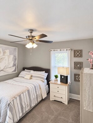 A bedroom with a bed, bedside table, lamp, ceiling fan, and artwork. -     YOUR Cozy 2brm Upper Kenmore Retreat w balcony near Buffalo & Niagara Falls. (Kenmore)