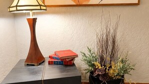 Huoneessa on moderni lamppu, geometrinen abstrakti maalaus, pieni sivupöytä, jossa on kirjoja, sekä koristeellinen kasvi kuviollisessa ruukussa.