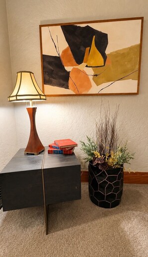 Una habitación con una lámpara moderna, un cuadro abstracto geométrico, una mesita de noche con libros y una planta decorativa en una maceta estampada.