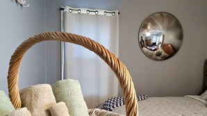 Huoneessa on punottu tuoli, kuviollinen vuodevaatteilla varustettu sänky, pyöreä peili ja verho.