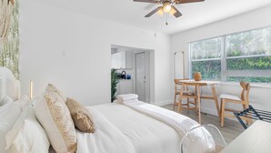 Een slaapkamer met een bed, een plafondventilator, een raam met zonwering en uitzicht op een keuken.