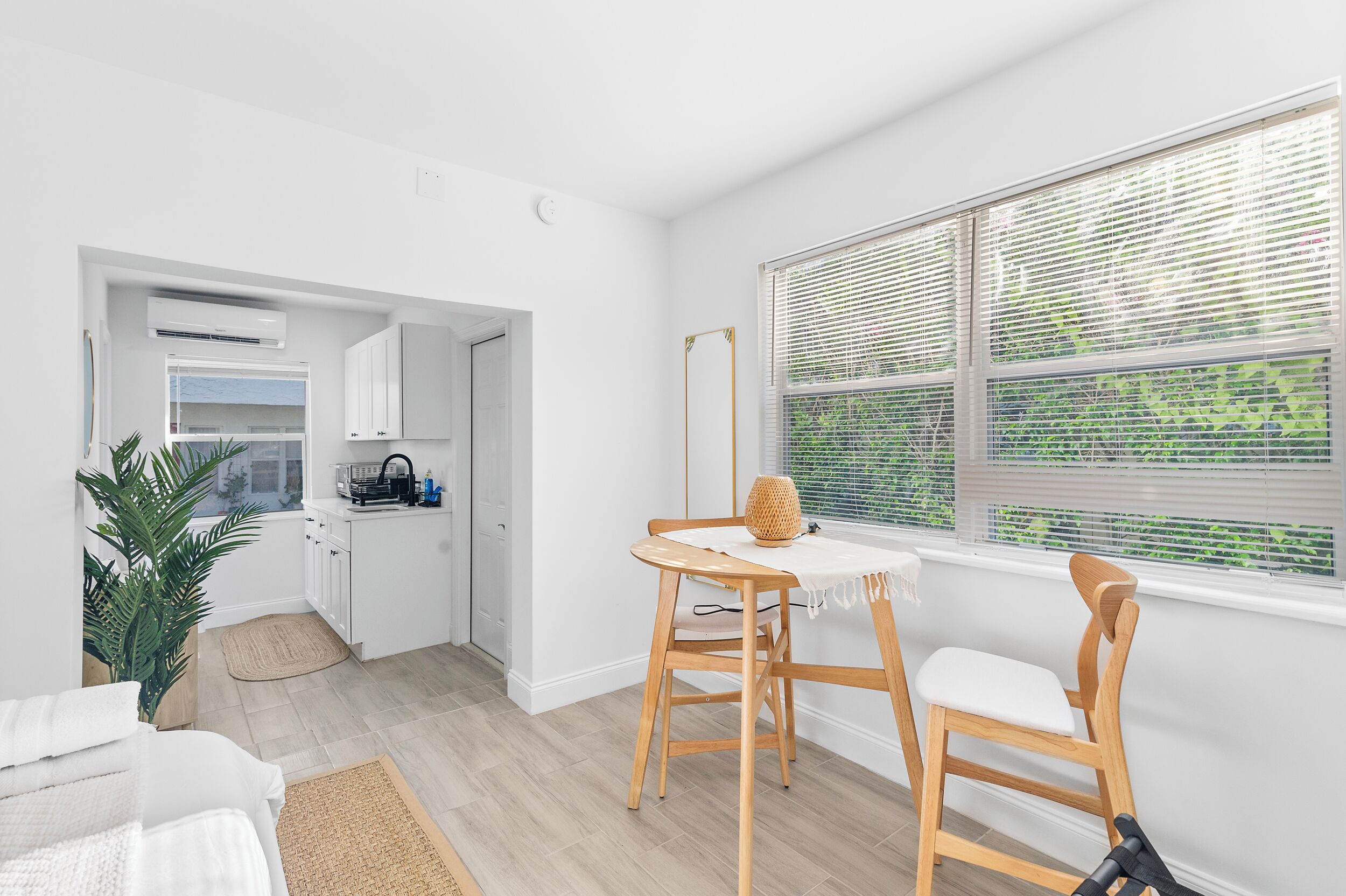 Une pièce moderne et minimaliste, avec une table à manger en bois, deux chaises, une grande fenêtre avec stores, un petit coin cuisine et une plante en pot.