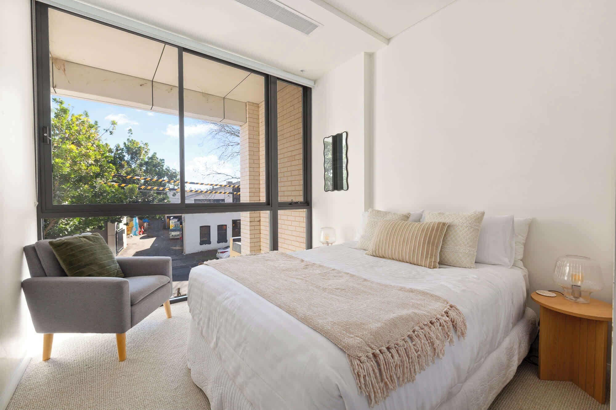 A bedroom with a large window, a bed with multiple pillows, a chair, and a bedside table with a lamp.