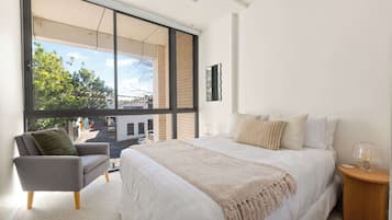 A bedroom with a large window, a bed with multiple pillows, a chair, and a bedside table with a lamp.