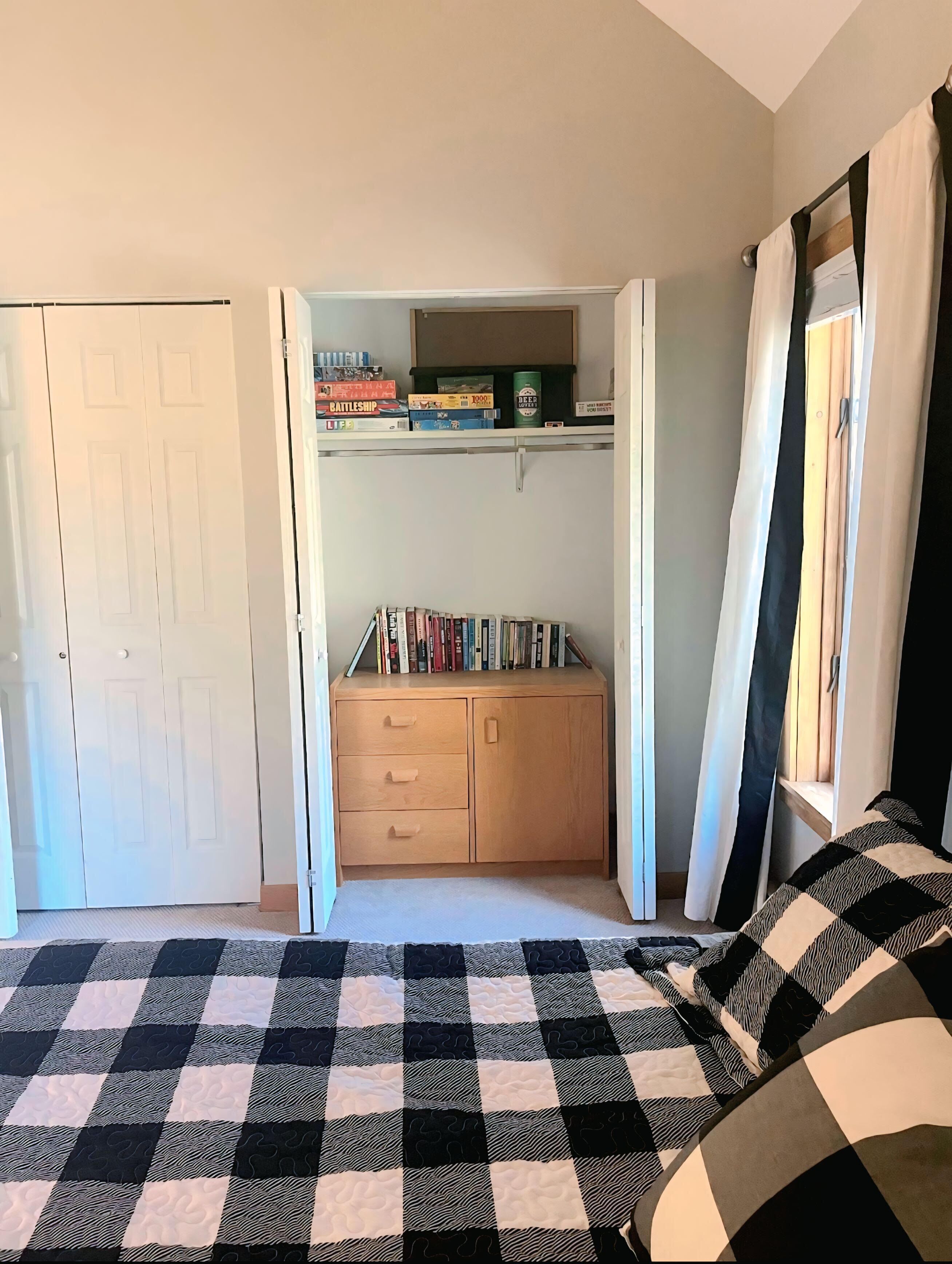 Un dormitorio con cama, armario, estante con libros y juegos, y ventana con cortinas.