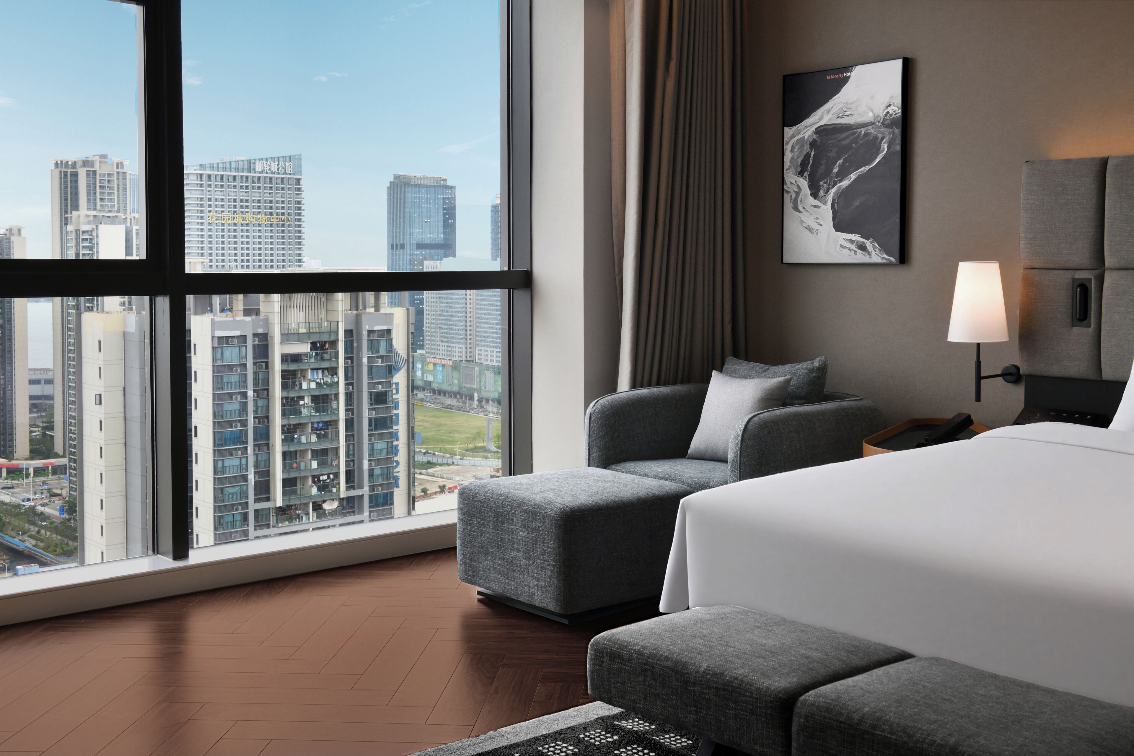A modern hotel room with a large window, a bed, a grey armchair, a small ottoman, a nightstand, and a lamp.