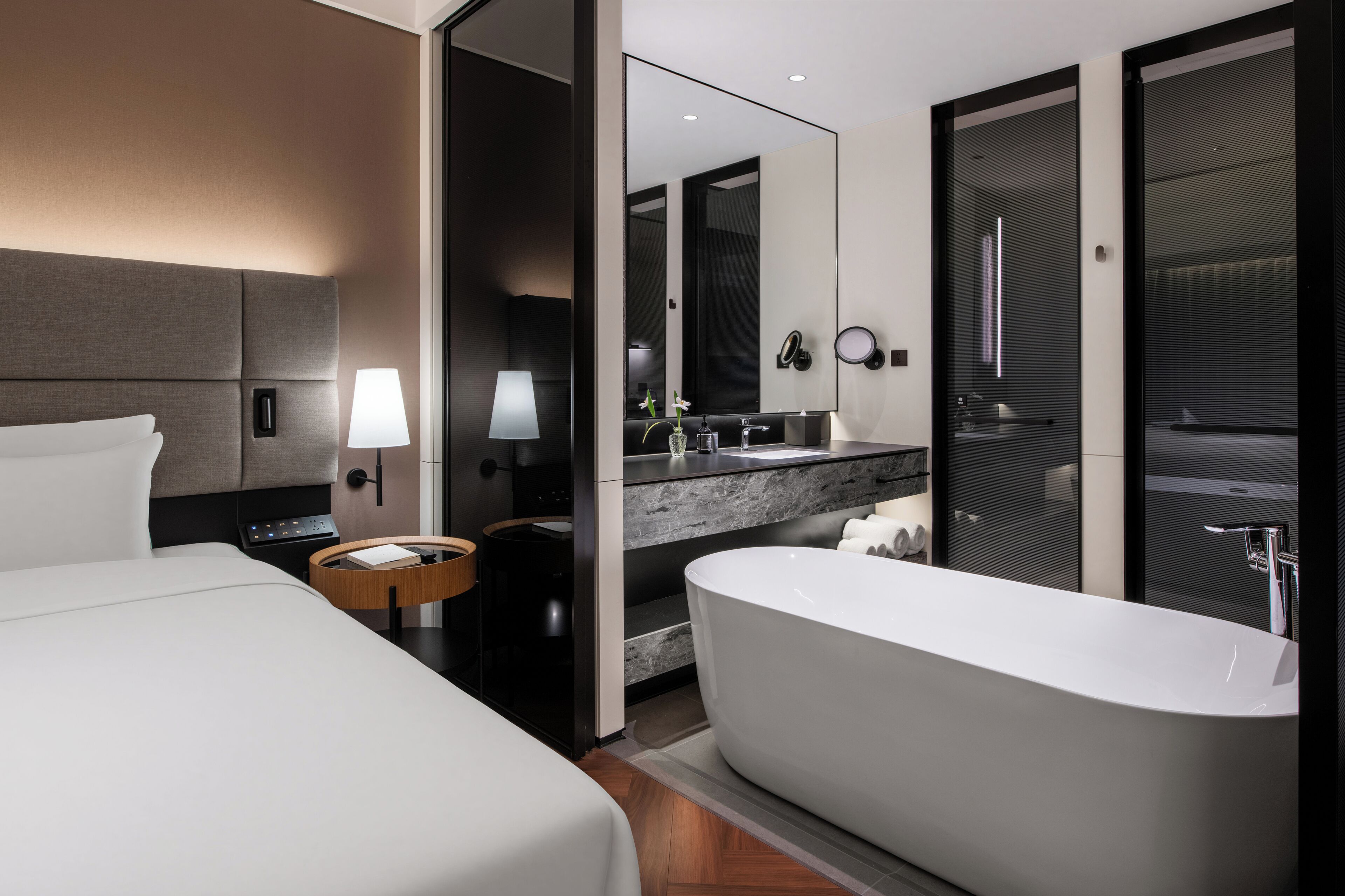 A modern hotel room with a large bed, a separate bathroom with a freestanding bathtub, a vanity with a mirror, and a nightstand with a lamp.