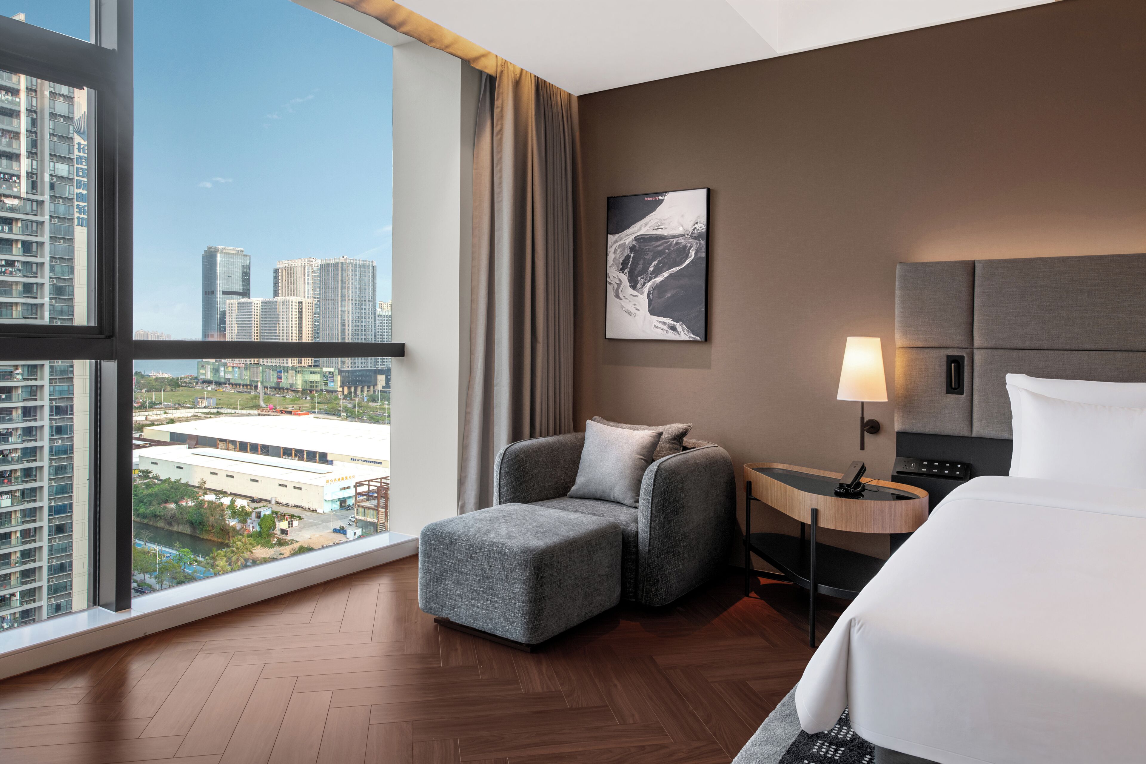 A hotel room with a large window, a bed, a chair, a nightstand, and a lamp.