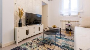 A modern living room with a flat-screen TV, white cabinetry, a small round table, a colorful rug, and a window with curtains. - Atocha VI (Madrid)
