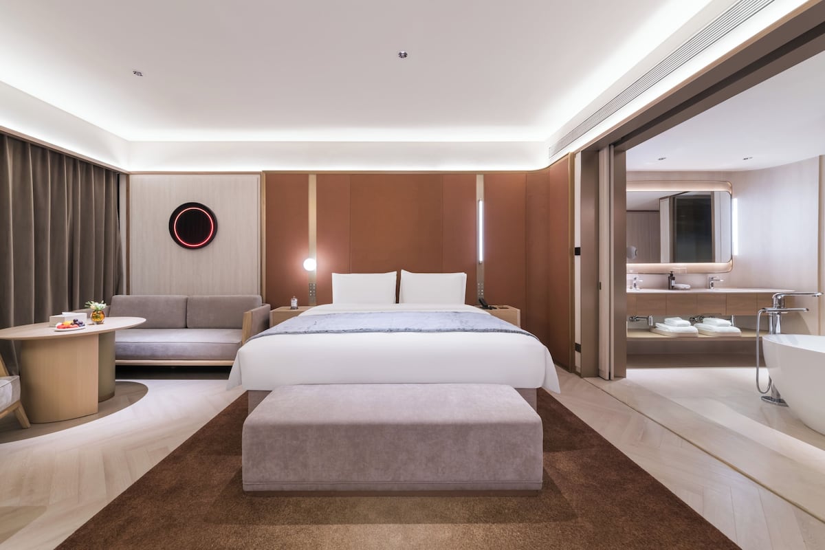 Una habitación de hotel moderna con cama, sofá, una mesita, un espejo redondo y un baño con bañera.