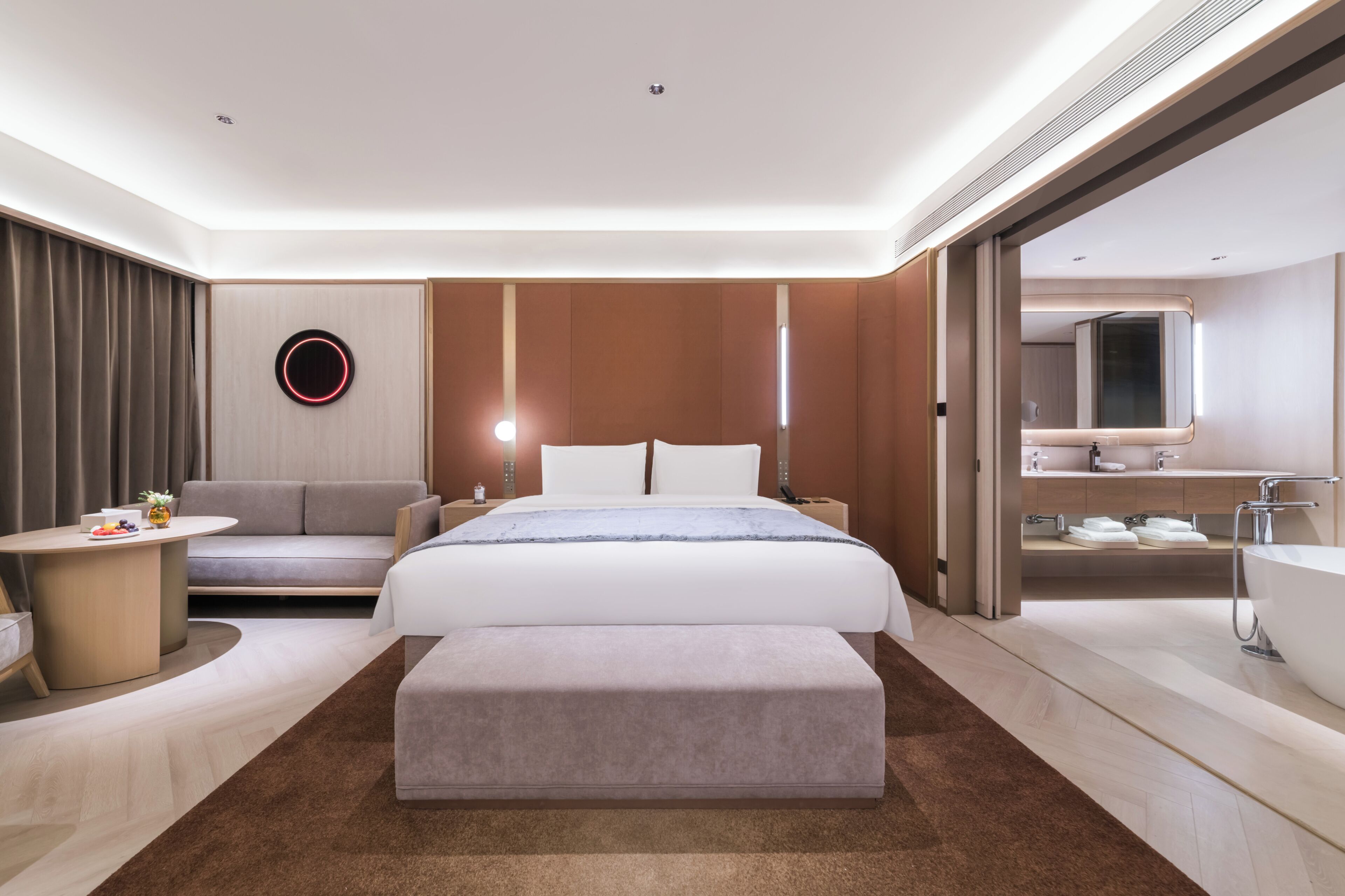 Moderni hotellihuone, jossa on sänky, sohva, pieni pöytä, pyöreä peili ja kylpyammeella varustettu kylpyhuone.