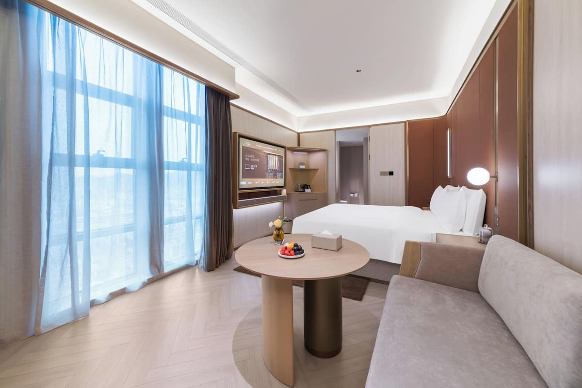 Habitación de hotel con cama, una mesa redonda con un florero y fruta, un sofá, un televisor y un ventanal con cortinas.