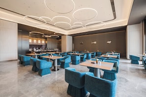 A modern restaurant with blue upholstered chairs and tables, circular ceiling lights, and a visible kitchen area.