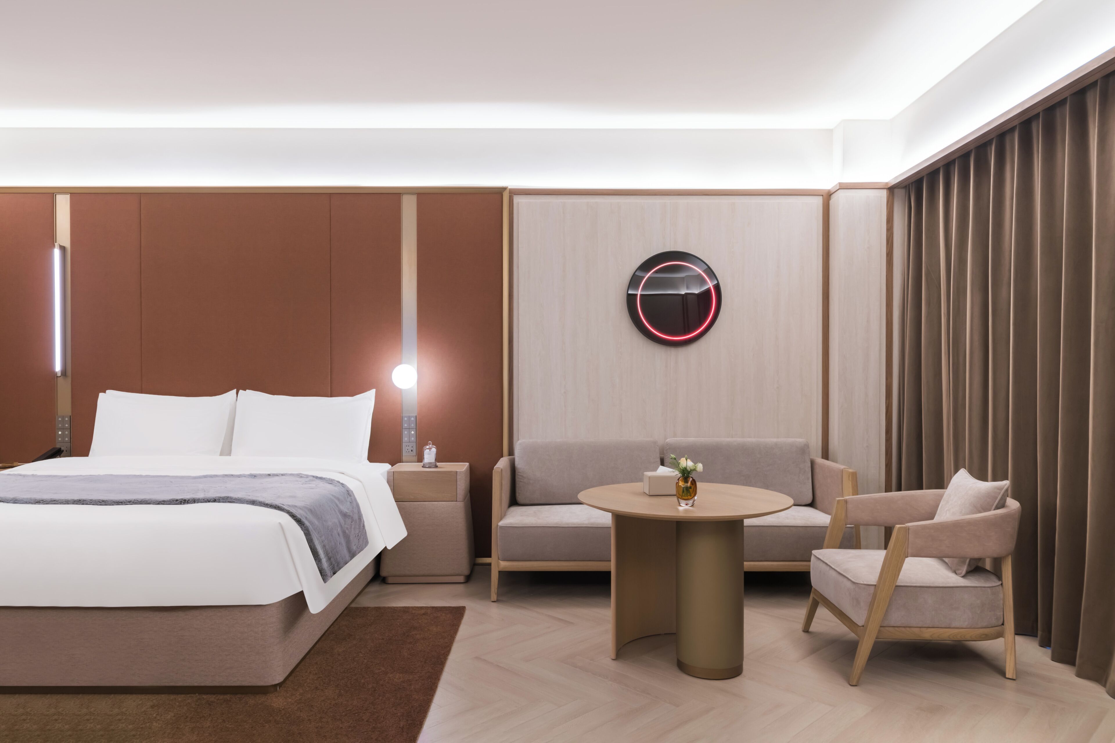 A modern hotel room with a bed, a sofa, a chair, a round table, and a wall-mounted light fixture.