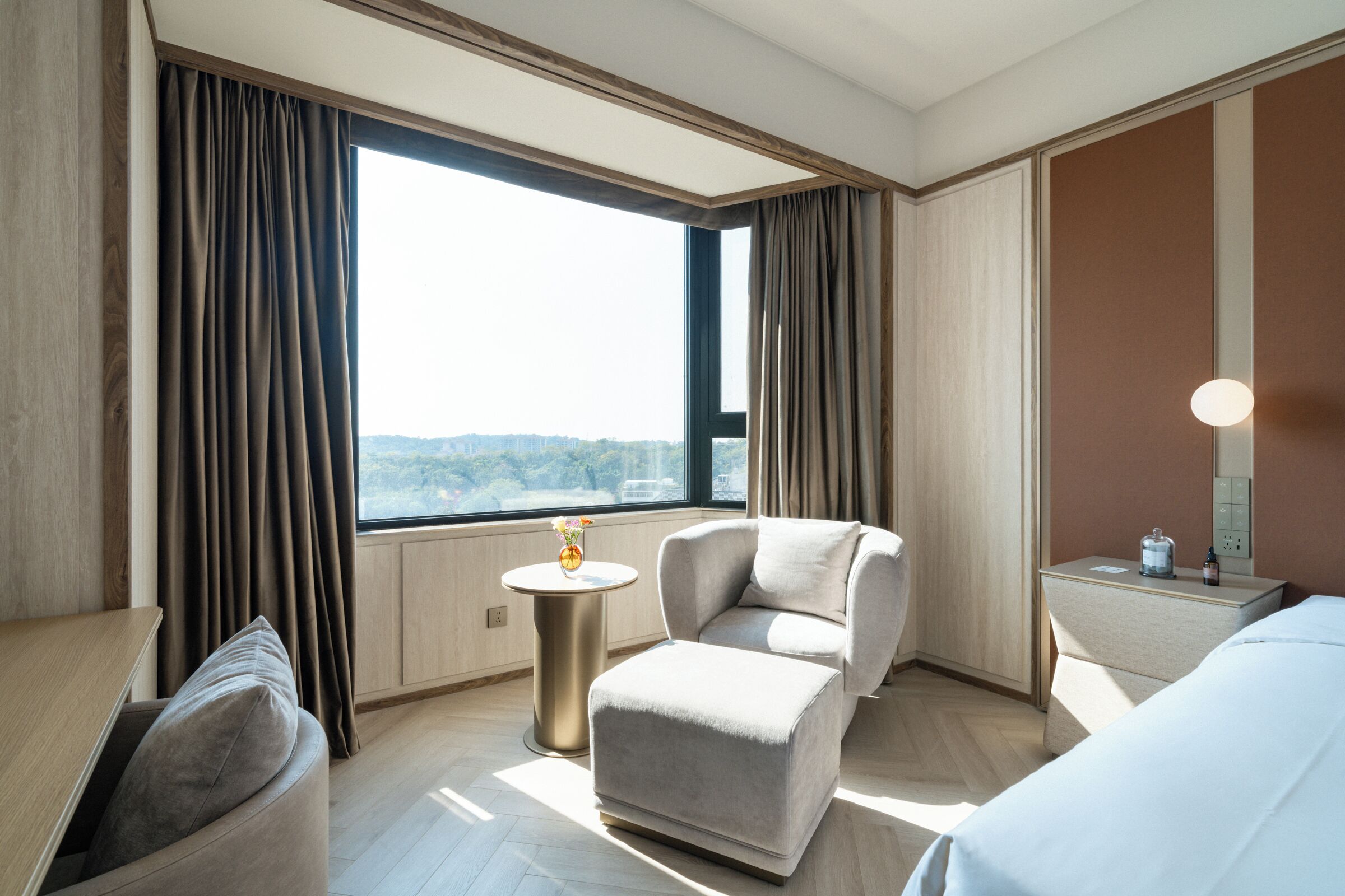 A hotel room with a large window, a round table with a vase, two armchairs, a bedside table with a lamp, and a bed.