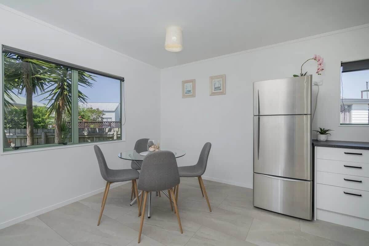 Una cucina moderna con un frigorifero in acciaio inox, una zona pranzo con tavolo in vetro e sedie grigie, e una finestra con vista su palme.