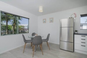 Una cocina moderna con un refrigerador de acero inoxidable, un comedor con una mesa de vidrio y sillas grises, y un ventanal con vista a palmeras.