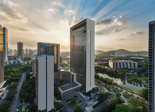 Un hotel moderno de altura, con un diseño vertical llamativo, rodeado de un paisaje urbano cuidado y un río.