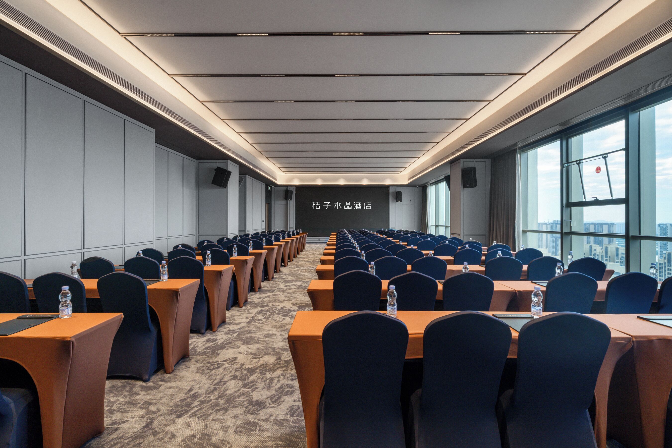 A conference room with rows of tables and chairs, a carpeted floor, and a city view through large windows.