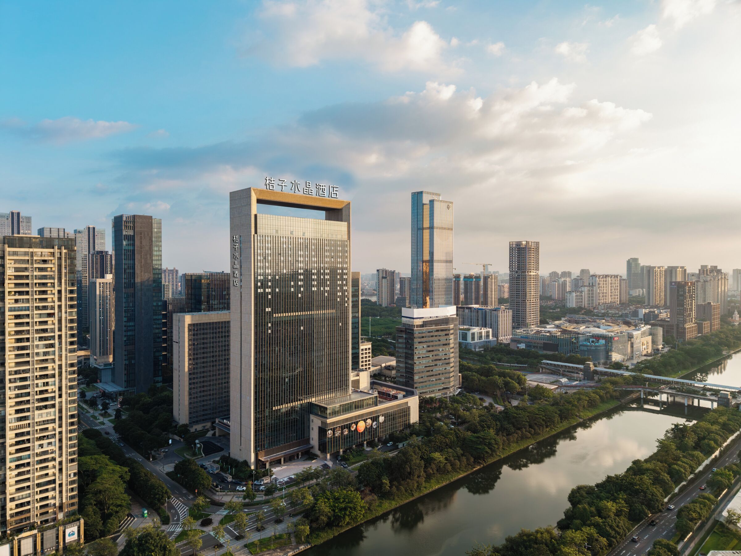 A high-rise hotel with a modern glass facade, situated next to a river with a bridge in the background.