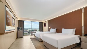 Quarto de hotel com duas camas, uma escrivaninha, televisão e uma janela grande com vista para a cidade.