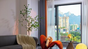 A modern living room with a sofa, a unique orange chair, a potted plant, a vase with flowers, and a view of a cityscape through a large window. - Local Vibe Downtown Guesthouse (Zhangjiajie)