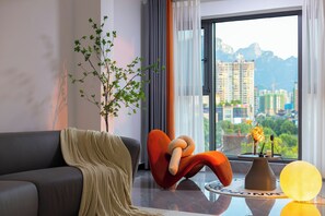 A modern living room with a sofa, a unique orange chair, a potted plant, a vase with flowers, and a view of a cityscape through a large window. - Local Vibe Downtown Guesthouse (Zhangjiajie)