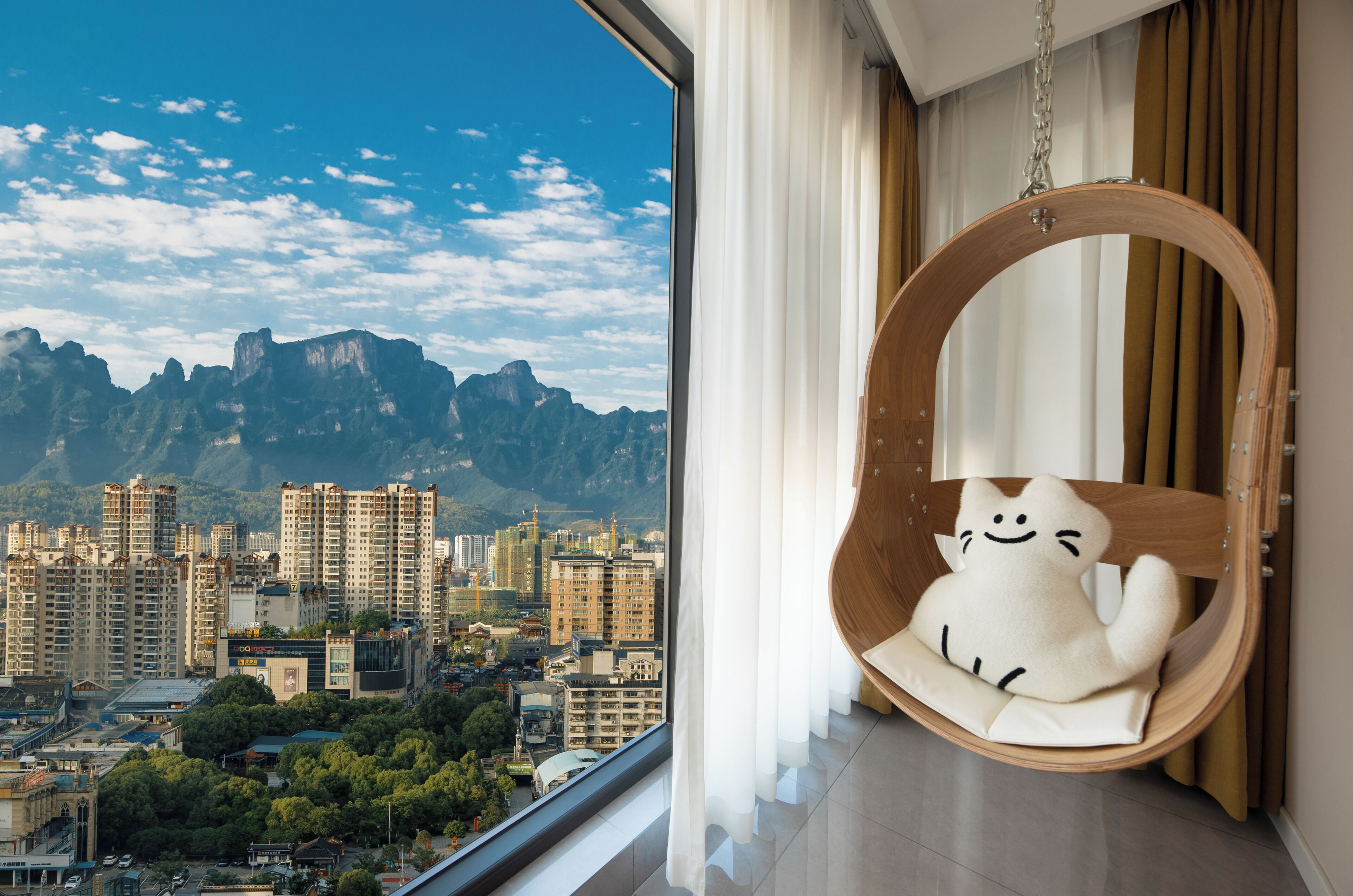 Een kamer met een groot raam dat uitzicht biedt op de stad, een houten schommelstoel met een pluche speeltje en een gordijn.
