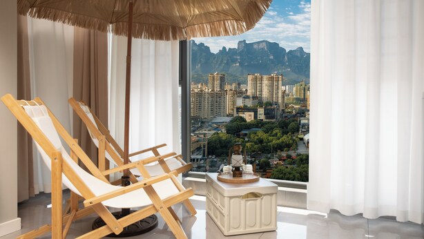 A balcony with two wooden chairs, a small table with a decorative item, and a view of a cityscape with mountains in the background. - Local Vibe Downtown Guesthouse (Zhangjiajie)