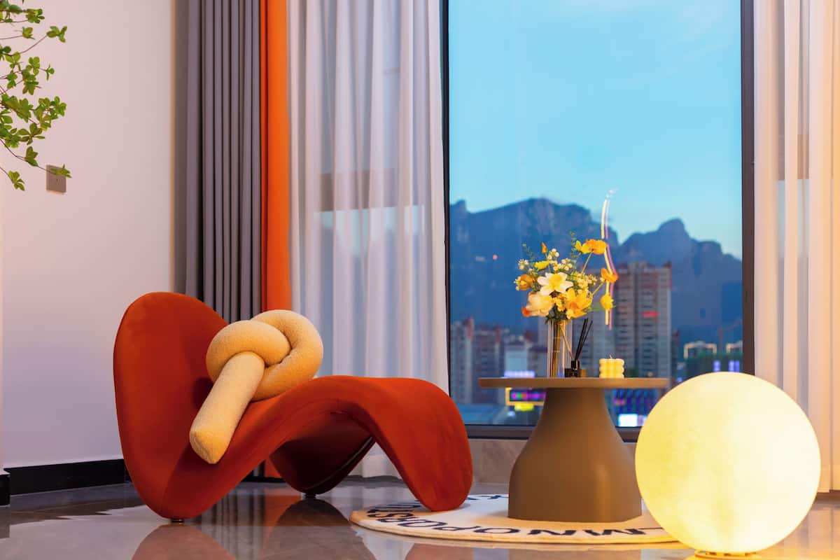 A modern room with a large window, a red chair, a round table with a vase of flowers, and a spherical light fixture.