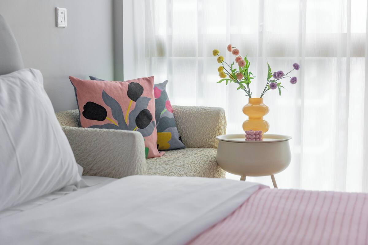 A bedroom with a bed, a sofa with colorful cushions, a small side table with a vase of flowers, and sheer curtains.