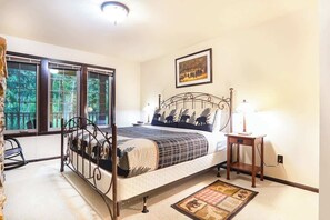 Un dormitorio con cama, una mesita de noche con lámpara, una ventana con vista a árboles y un cuadro en la pared.