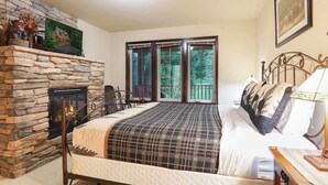 Ein Schlafzimmer mit Kamin aus Stein, einem Bett mit kariertem Überwurf, einem Nachttisch und einem Fenster mit Blick auf Grünflächen.