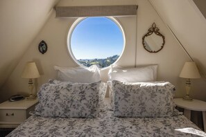 Ein Schlafzimmer mit einem Rundfenster, zwei Nachttischen mit Lampen, floralem Bettbezug und Blick auf eine Landschaft.