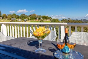 A table with a glass bowl of flowers, two wine glasses, and a bottle on a wooden deck with a white railing and a scenic view. - Garden and Bird Haven (Waiheke Island)