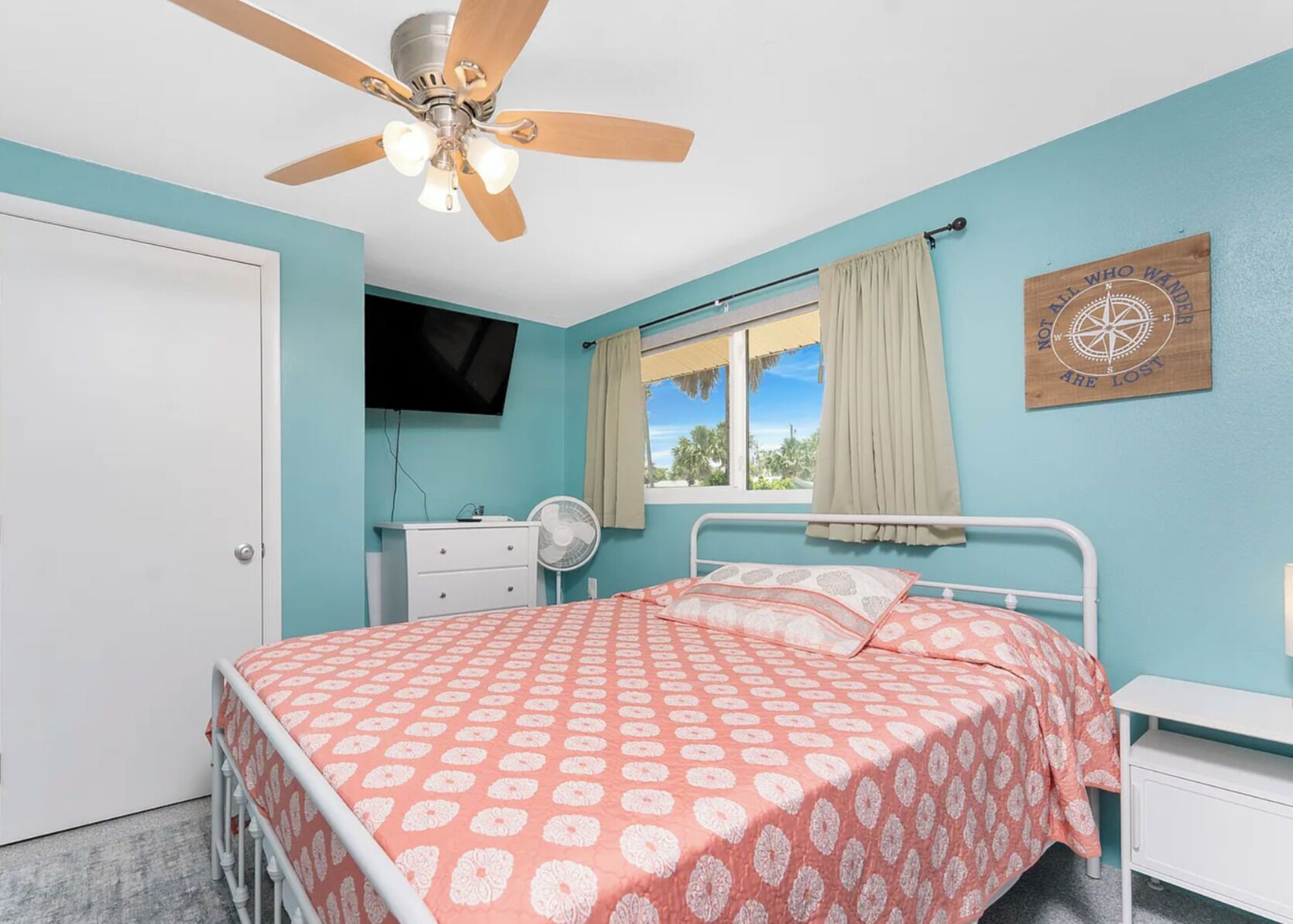 A bedroom with a bed, a ceiling fan, a window with curtains, a TV, and a wall decoration.