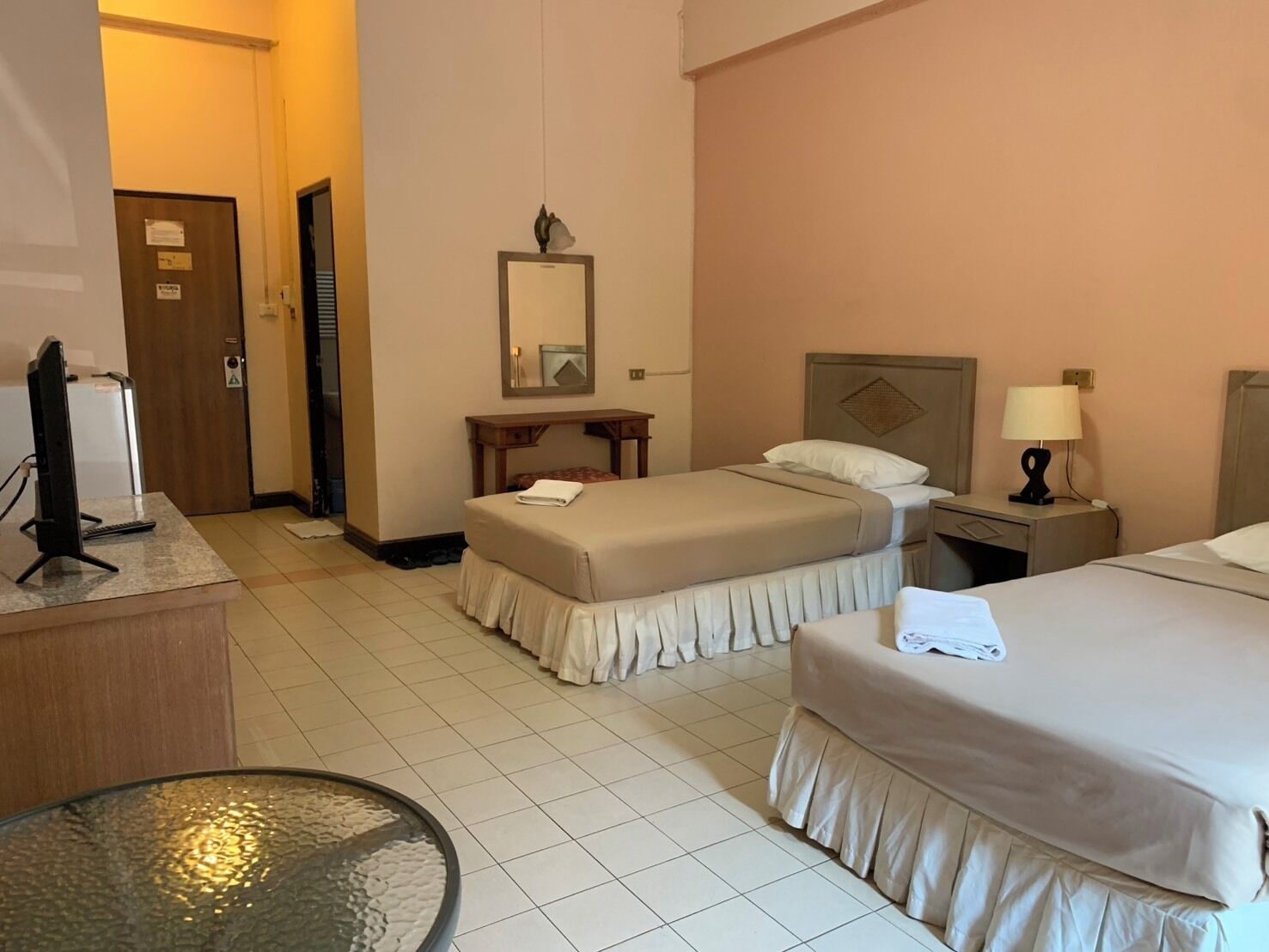 A hotel room with two beds, a desk, a chair, a round table, a mirror, and a door.